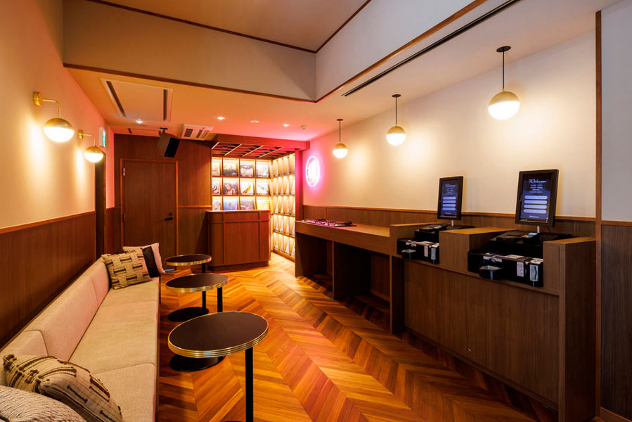 1F フロント：店舗のようなフロントで常設の飾り棚よりレコードをリクエストして、かけて頂く事が可能です。オーナーが音響にも拘り、設備を整えてお出迎えいたします。ヘリンボーンの無垢材、落ち着いた木目が高級感を与えています。自動チェックイン機も備え、予約をしていれば待たずにチェックインOKです。