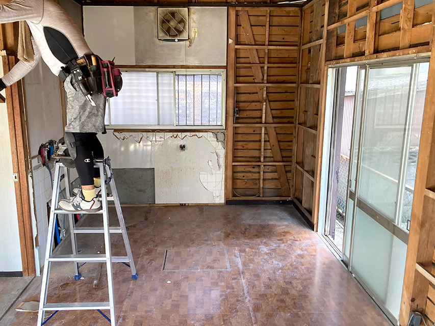 キッチン周りの壁材を撤去し、下地が見える室内の様子。作業員が脚立で撤去作業を進めており、床下点検口の位置も確認できる写真