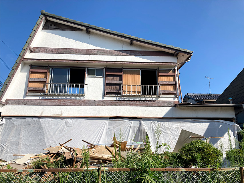 戸建住宅の外観を正面から写した写真で、足場用シートがかけられ解体材が積まれている様子から、外壁工事前の状況を説明しています。